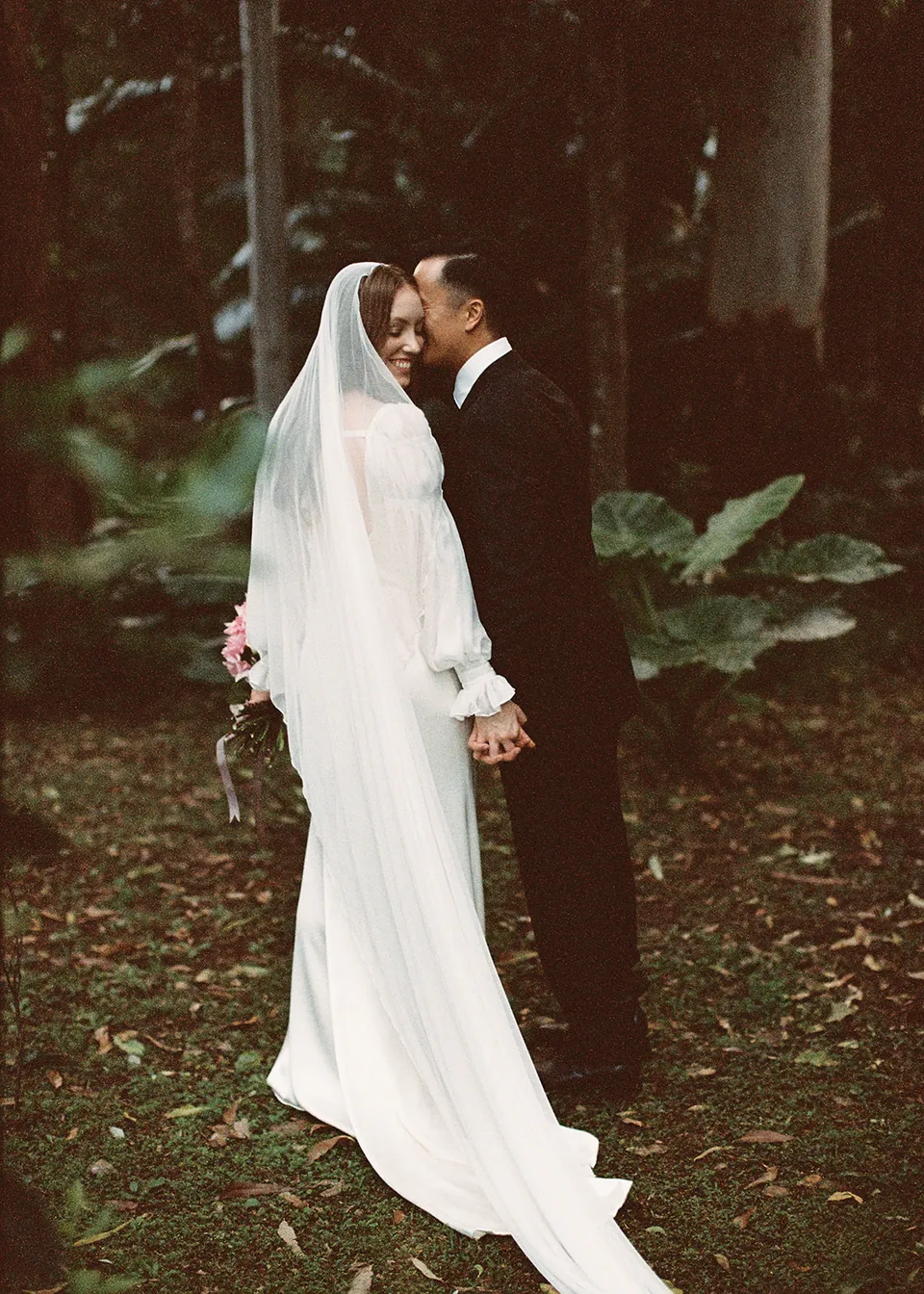 On 35mm film: Elyse and Jonathan standing close together beneath the garden trees. At Fig Tree Villa in Ewingsdale, Byron Bay, for an Elopement Collective elopement. Photograph by Bulb Creative.