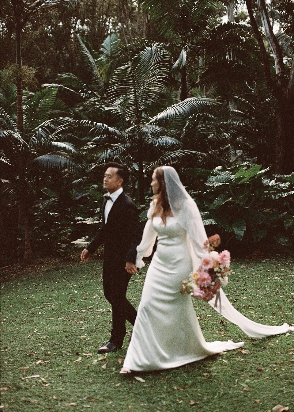 On 35mm film: The couple walking hand in hand past the tree ferns. At Fig Tree Villa in Ewingsdale, Byron Bay, for an Elopement Collective elopement. Photograph by Bulb Creative.