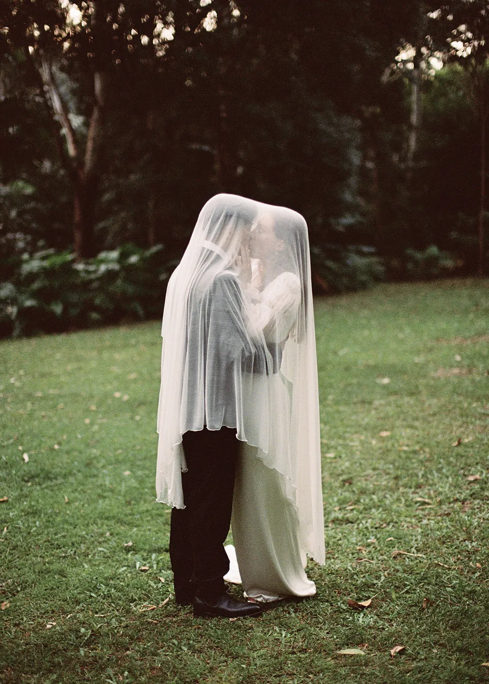 On 35mm film: Full-length portrait of the couple standing together beneath Elyse's veil. At Fig Tree Villa in Ewingsdale, Byron Bay, for an Elopement Collective elopement. Photograph by Bulb Creative.