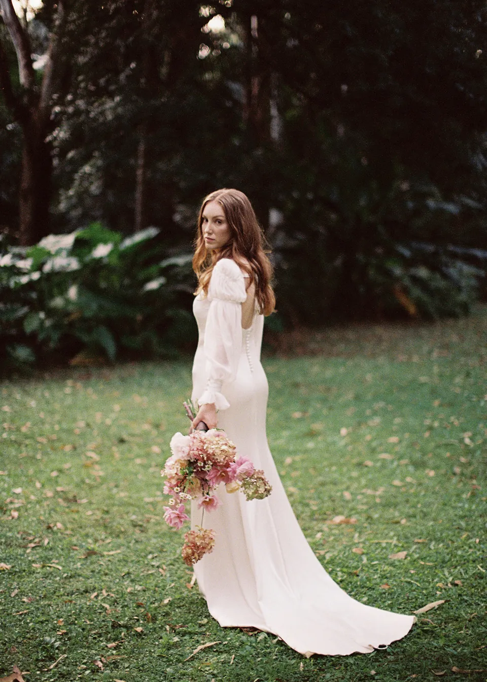 On 35mm film: Elyse looking back with her long dress trailing across the lawn. At Fig Tree Villa in Ewingsdale, Byron Bay, for an Elopement Collective elopement. Photograph by Bulb Creative.