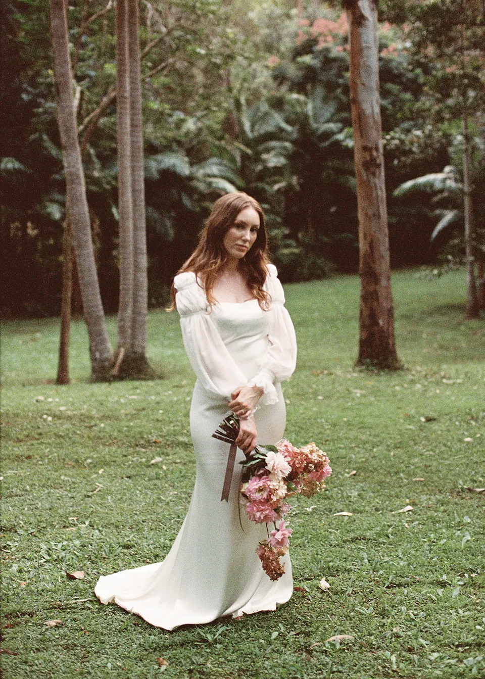 On 35mm film: Elyse smiling with her bouquet at her side beneath tall trees, with flowers by The Perfect Party Co. At Fig Tree Villa in Ewingsdale, Byron Bay, for an Elopement Collective elopement. Photograph by Bulb Creative.