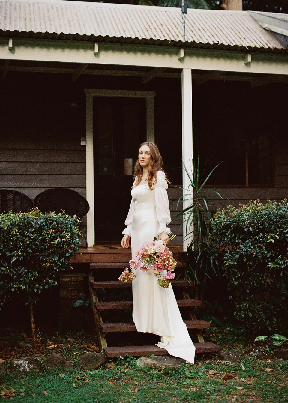 On 35mm film: Elyse standing beside the steps of the weatherboard cottage. At Fig Tree Villa in Ewingsdale, Byron Bay, for an Elopement Collective elopement. Photograph by Bulb Creative.