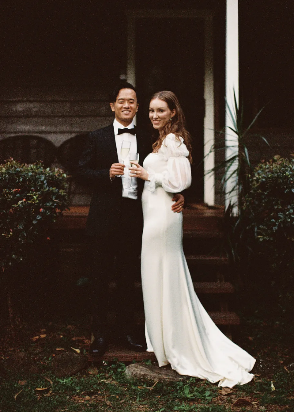 On 35mm film: Elyse and Jonathan smiling together with glasses in hand outside the cottage. At Fig Tree Villa in Ewingsdale, Byron Bay, for an Elopement Collective elopement. Photograph by Bulb Creative.