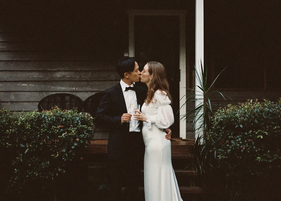 On 35mm film: The couple sharing a kiss beside the verandah while holding their glasses. At Fig Tree Villa in Ewingsdale, Byron Bay, for an Elopement Collective elopement. Photograph by Bulb Creative.
