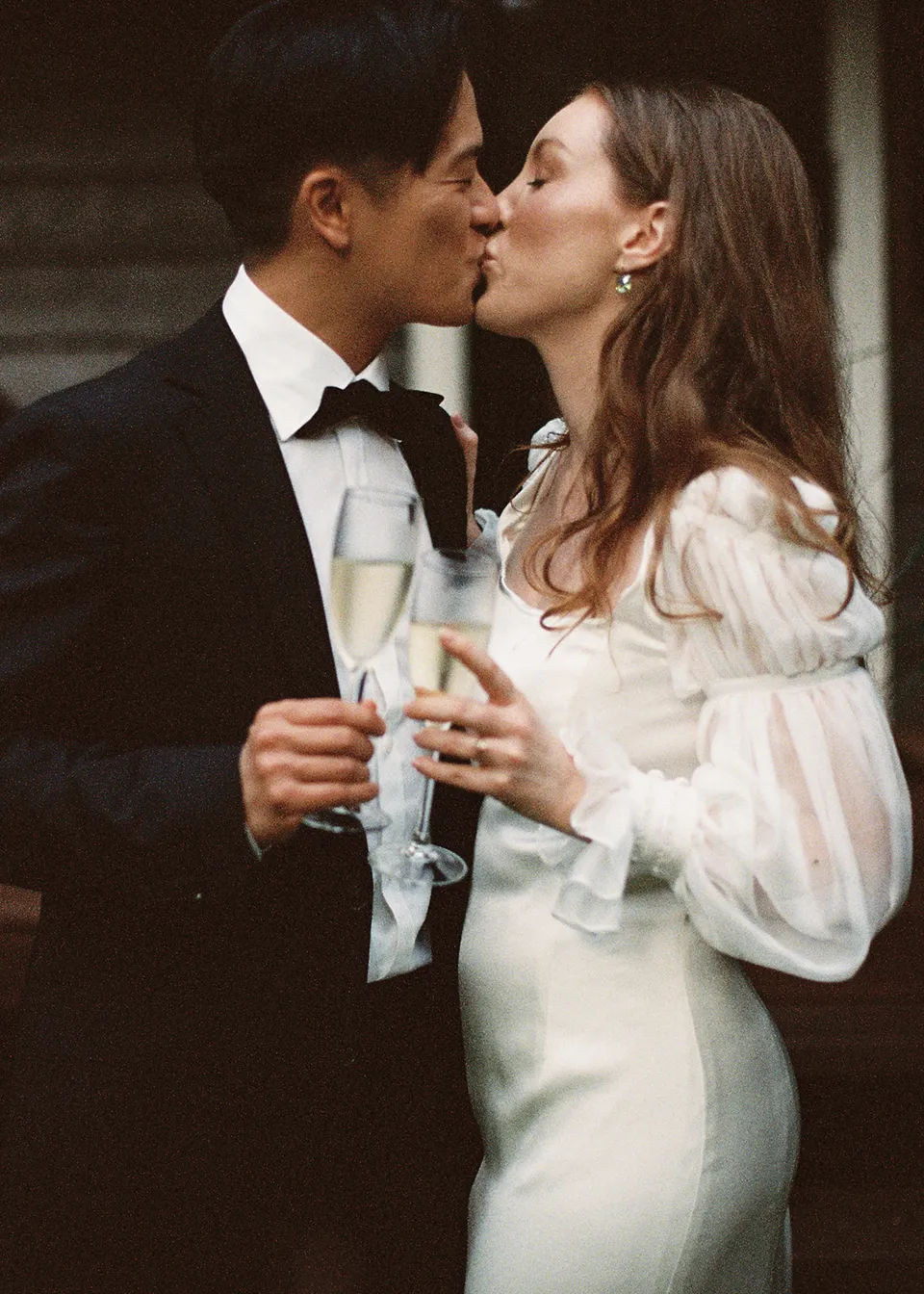 On 35mm film: Elyse and Jonathan kissing with their glasses raised between them. At Fig Tree Villa in Ewingsdale, Byron Bay, for an Elopement Collective elopement. Photograph by Bulb Creative.