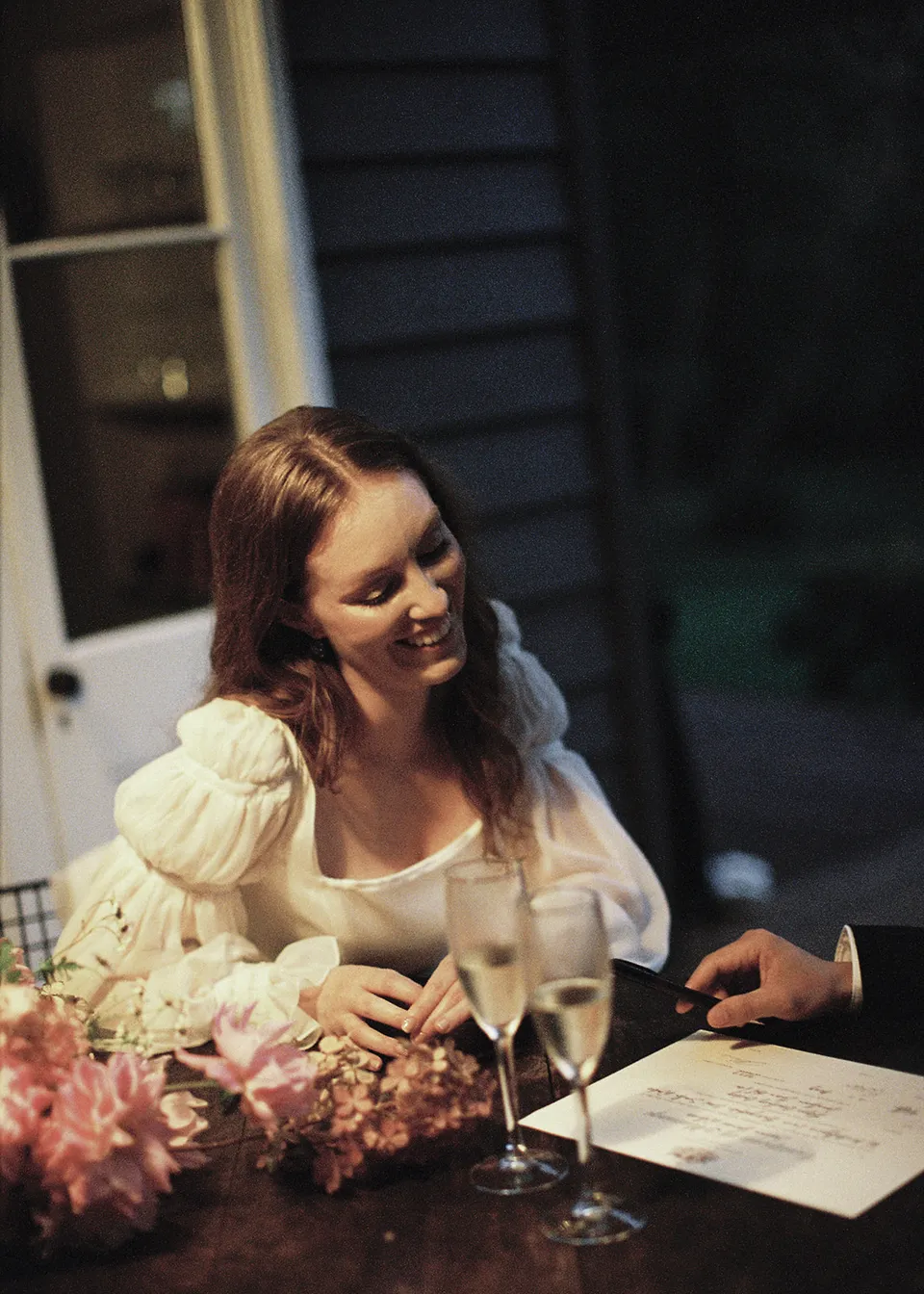 On 35mm film: Elyse smiling at the timber table with flowers and paperwork in front of her. At Fig Tree Villa in Ewingsdale, Byron Bay, for an Elopement Collective elopement. Photograph by Bulb Creative.