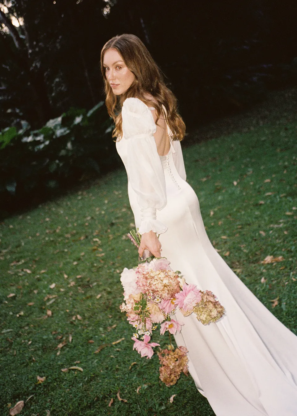 On 35mm film: Elyse looking back with her bouquet and long veil in the garden, with flowers by The Perfect Party Co. At Fig Tree Villa in Ewingsdale, Byron Bay, for an Elopement Collective elopement. Photograph by Bulb Creative.