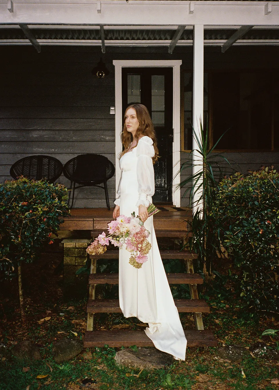 On 35mm film: Elyse standing beside the cottage steps with flowers at her side. At Fig Tree Villa in Ewingsdale, Byron Bay, for an Elopement Collective elopement. Photograph by Bulb Creative.