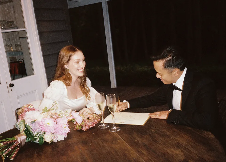 On 35mm film: Elyse and Jonathan laughing together beside their flowers and ceremony paperwork. At Fig Tree Villa in Ewingsdale, Byron Bay, for an Elopement Collective elopement. Photograph by Bulb Creative.