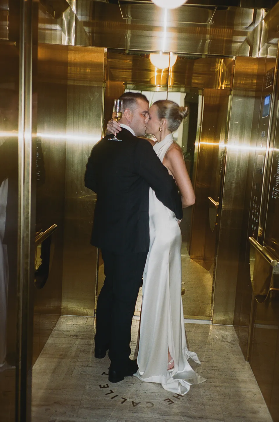 Clare and Matt kissing with champagne in a gold mirrored lift at The Calile Hotel, Fortitude Valley, during their Elopement Collective elopement in Brisbane.
