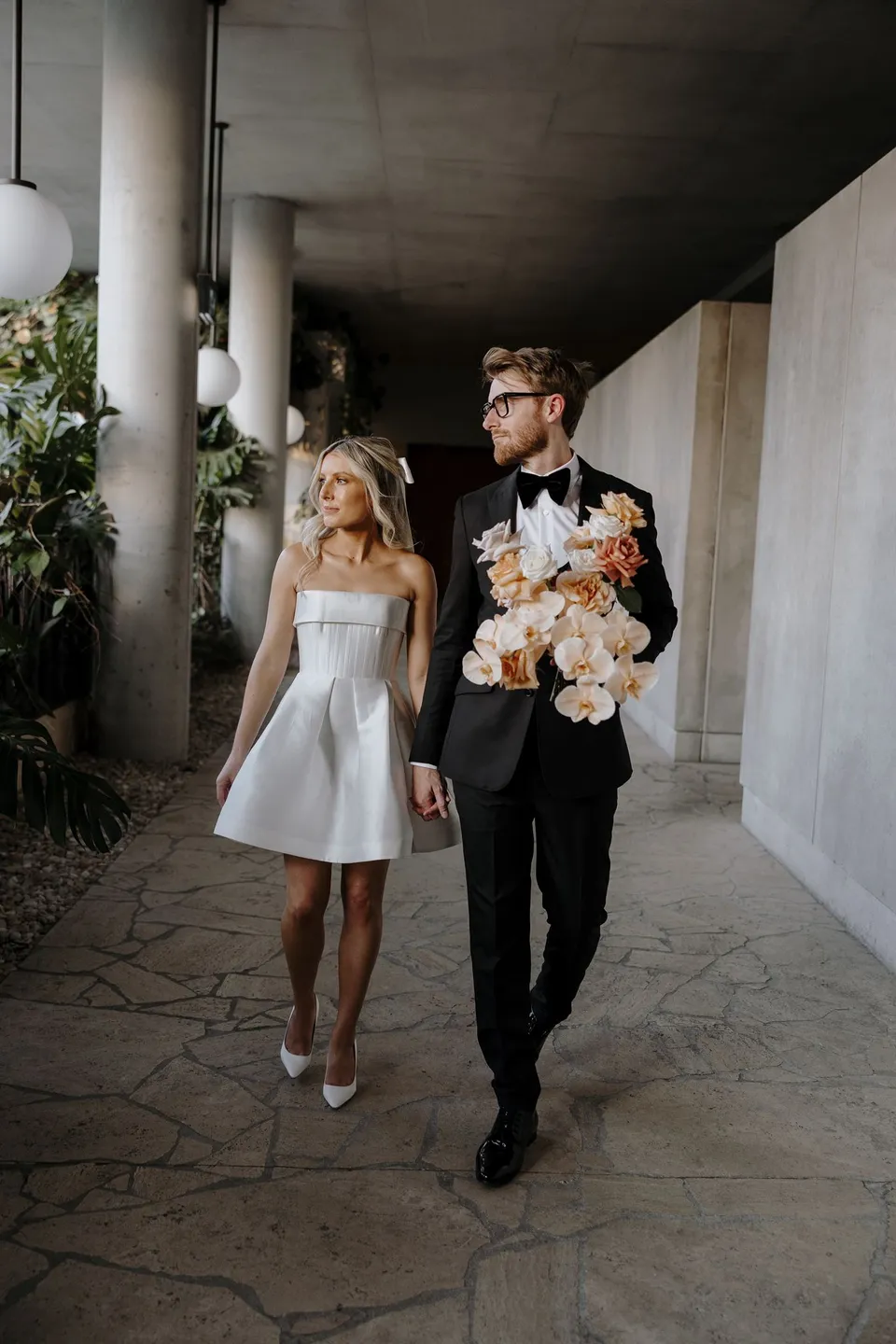 Danielle and Adam walking hand in hand beneath concrete columns, with Adam carrying the peach and white bouquet, during Danielle and Adam’s Elopement Collective elopement at The Calile Hotel in Fortitude Valley, Brisbane.