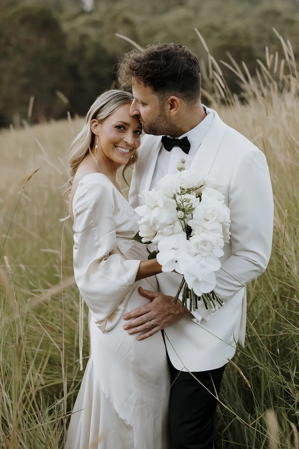 Greg kissing Gabrielle’s forehead as she smiles and holds her white bouquet in the paddock at Roundhill Retreat in Obi Obi, Sunshine Coast Hinterland, for their Elopement Collective elopement.