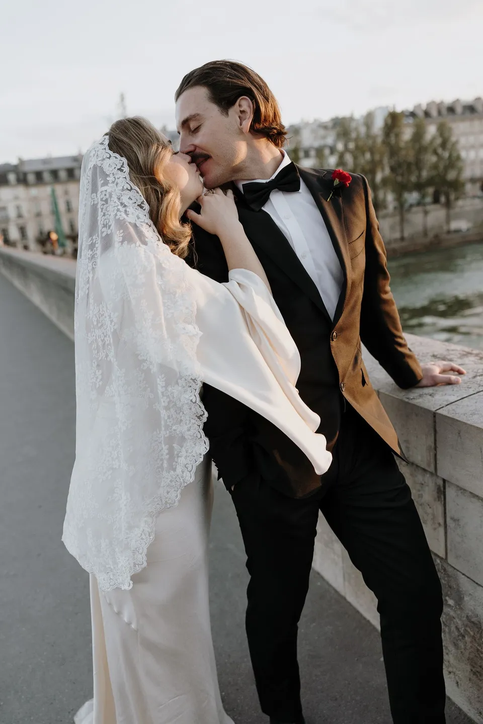 Kara and Samuel kissing beside a stone bridge balustrade above the Seine, with her lace veil, his red Ivy Bleu buttonhole and Parisian rooftops behind them, beside the Seine in Paris, France, during an Elopement Collective elopement. Photograph by George Bowden.