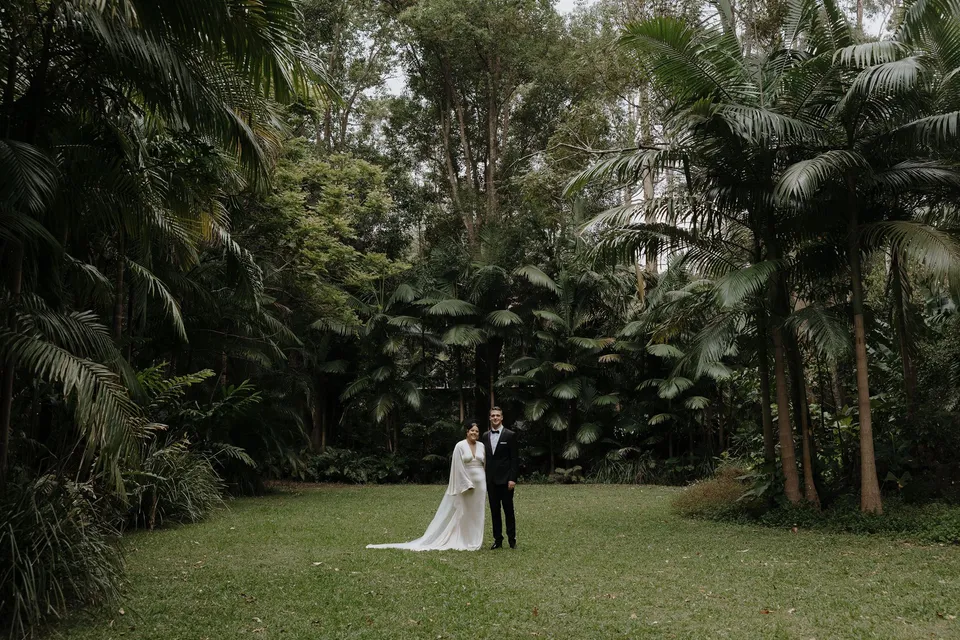 Laura and Matthew standing together on a lawn framed by tall palms and dense green foliage at Fig Tree Villa in Ewingsdale, Byron Bay. An Elopement Collective elopement, photographed by George Bowden.