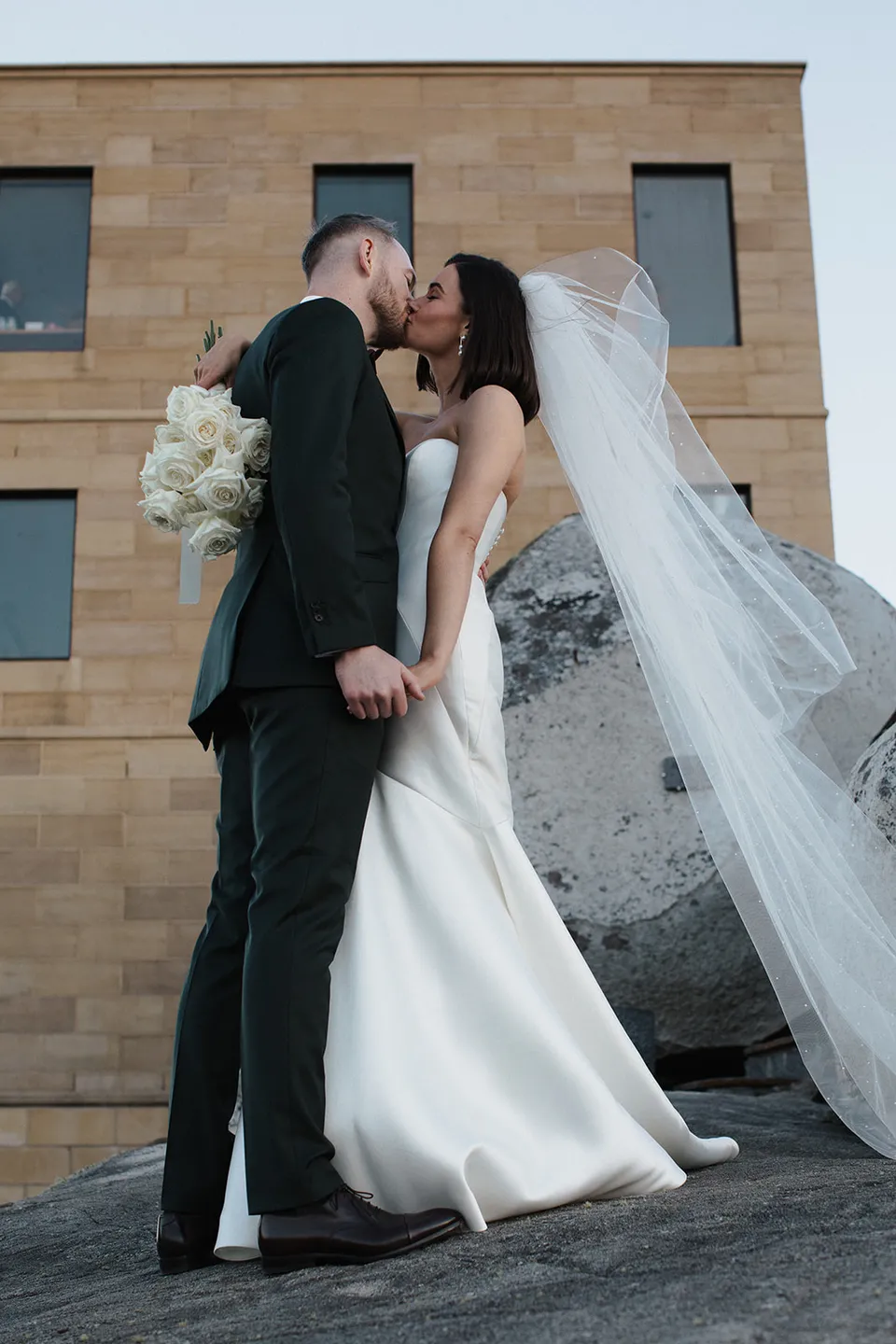 Meg and Ryan kissing beside the sandstone tower, with her veil lifting over a granite boulder and her white rose bouquet by The Romantics. At The Keep on Tasmania’s east coast, for an Elopement Collective elopement. Photograph by Jenna Agius.