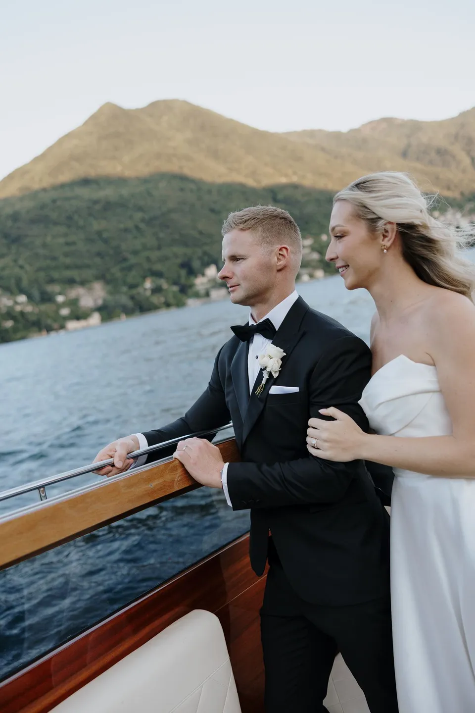 Zoe and Charlton standing beside the wooden rail of a boat, with mountains and lakeside homes behind them, on Lake Como in Lombardy, Italy, for an Elopement Collective elopement. Photo by George Bowden.