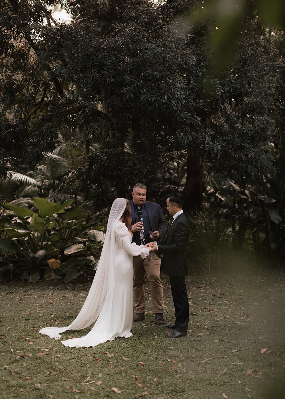The couple listening to celebrant Josh Withers during their garden ceremony. At Fig Tree Villa in Ewingsdale, Byron Bay, for an Elopement Collective elopement. Photograph by Bulb Creative.