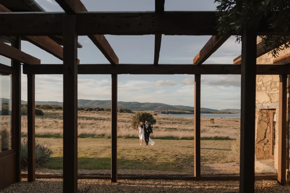 A couple walking towards a timber-framed stone courtyard, with coastal grassland and Great Oyster Bay beyond, at Thalia Haven on the East Coast of Tasmania, for an Elopement Collective elopement.