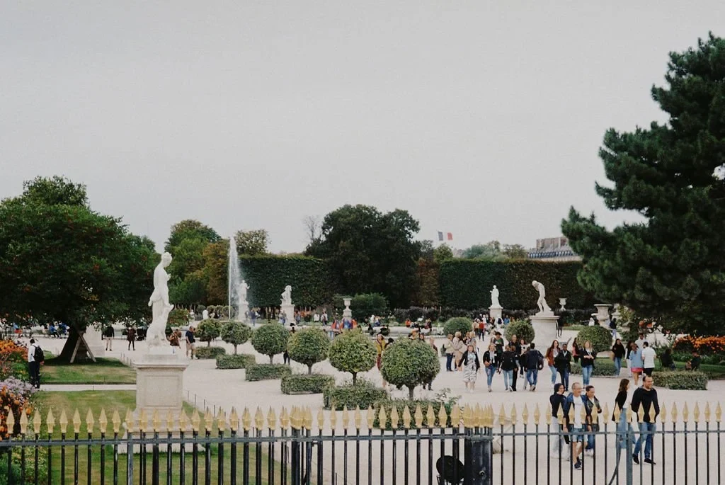 Park scene with statues, trees, people walking, and a fountain in the background.