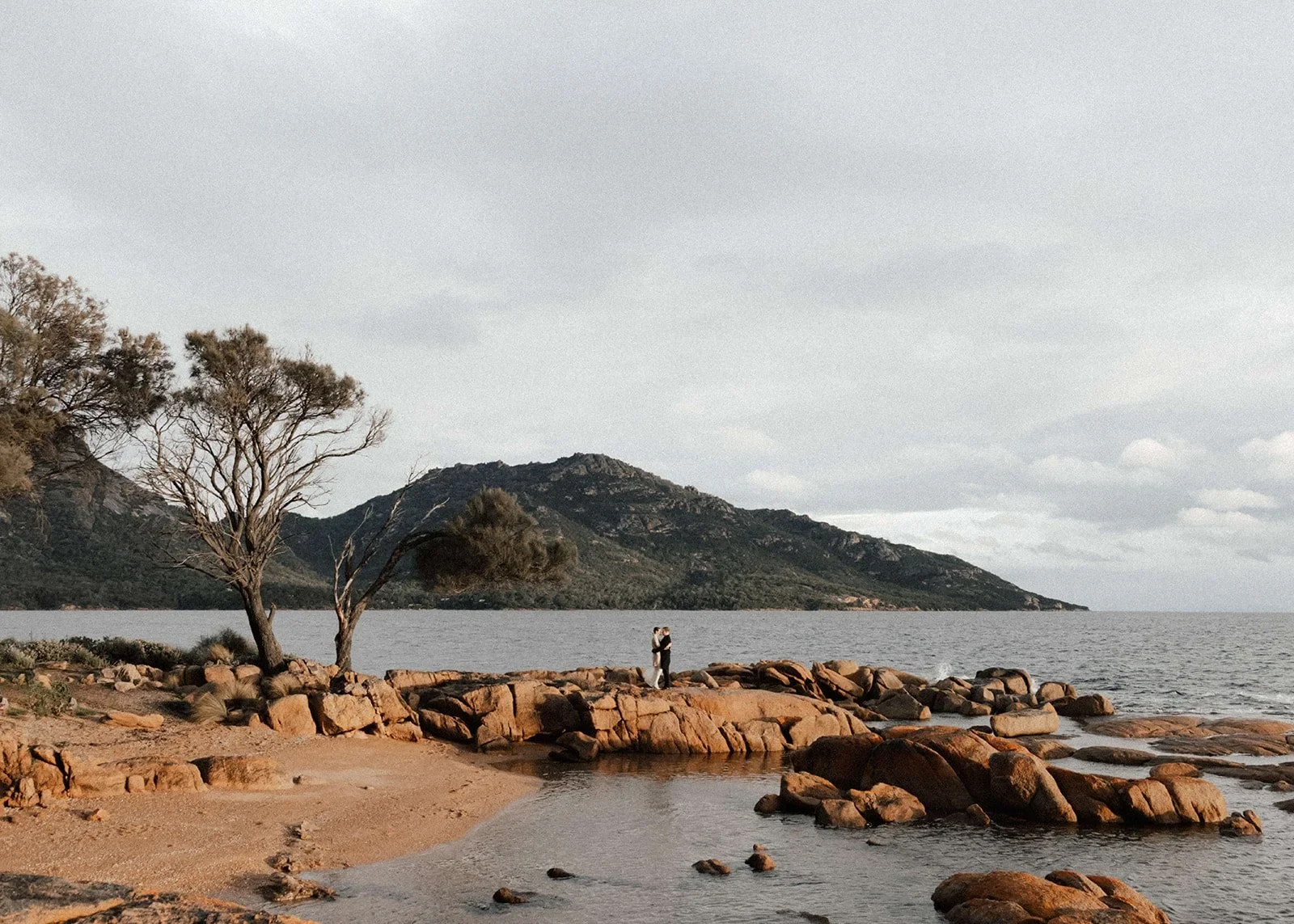 Couple standing together on a rocky shoreline with water and wooded hills behind them