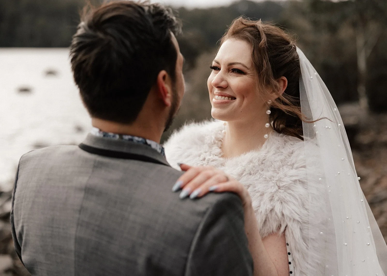 Sarah smiling at Jono with her hand resting on his shoulder and her veil gathered behind her, at Pumphouse Point, Lake St Clair, Tasmania, for an Elopement Collective elopement. Photo by Bulb Creative.