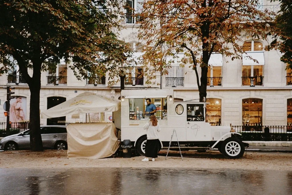 An ice cream truck in the style of a vintage car is parked on a street, with a person standing nearby. The truck is set up under a tarp, and there are trees and a building with large windows in the background. It's autumn, as evidenced by the fallen 