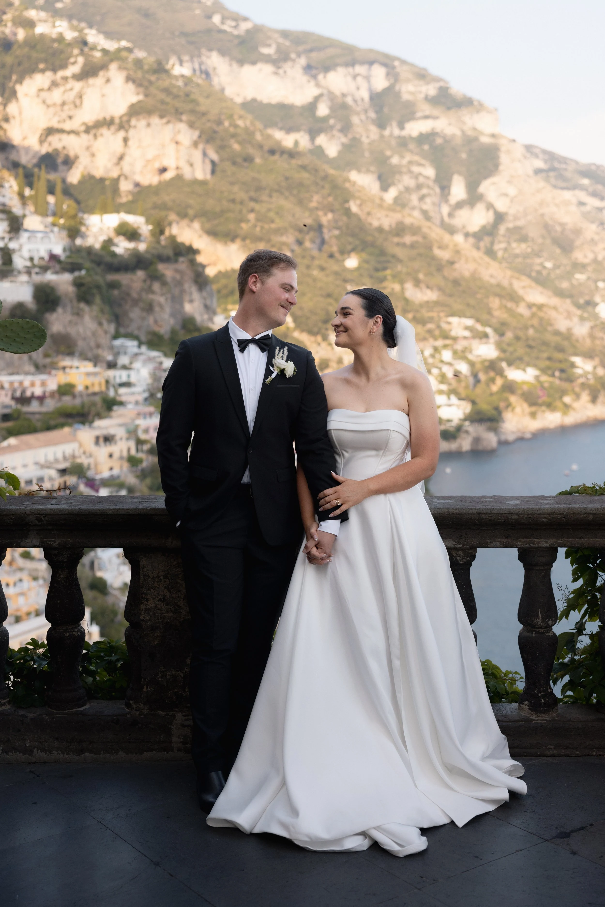 Couple in wedding attire holding hands on a terrace above a coastal landscape