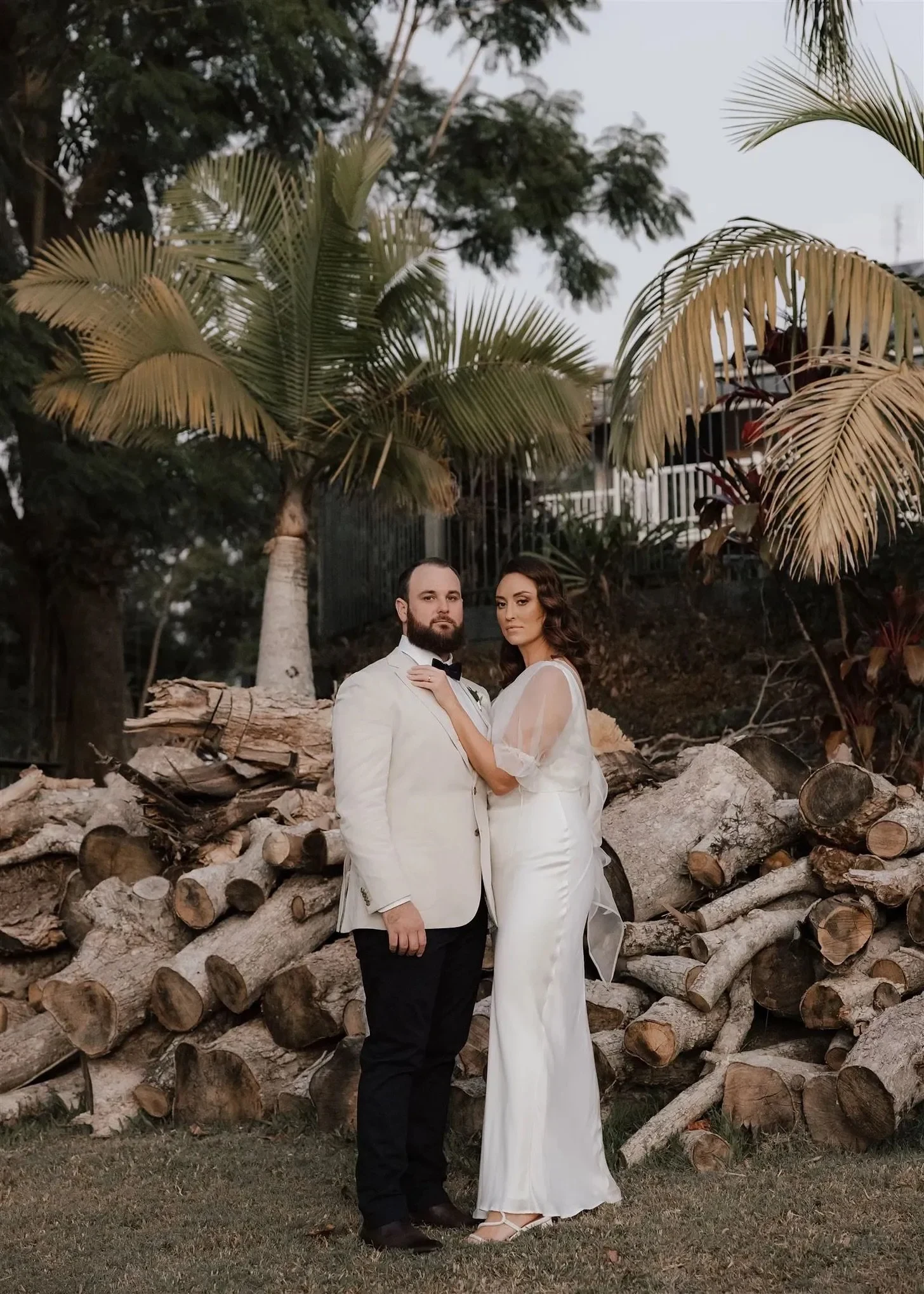 Tineeka and Chris standing together beside a stack of timber logs and tall palms, in Montville, Sunshine Coast Hinterland, Queensland, for an Elopement Collective elopement. Photo by Bulb Creative.