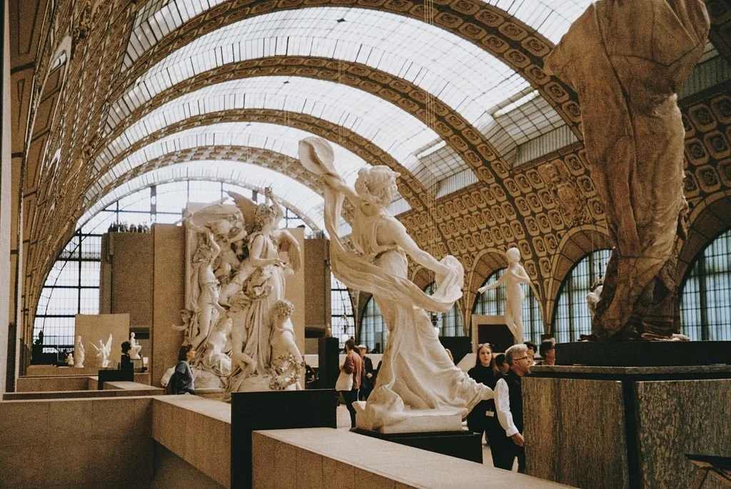 Interior of a museum with arched ceilings, featuring classical sculptures, including statues of figures in dynamic poses. Visitors walk among the art pieces, exploring the exhibit.