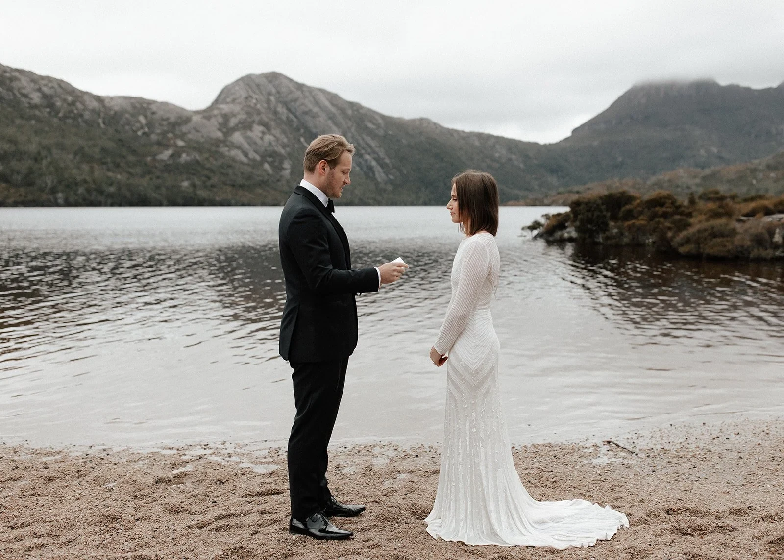 Josh reading from his vow booklet as April listens on the lakeshore, at Dove Lake, Cradle Mountain, Tasmania, for an Elopement Collective elopement. Photo by Bulb Creative.