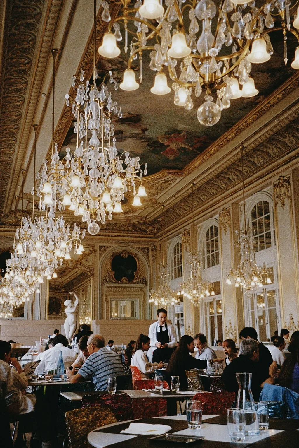 Luxurious restaurant interior with ornate chandeliers, detailed ceiling artwork, and diners seated at tables. A waiter is attending to customers in elegant surroundings.