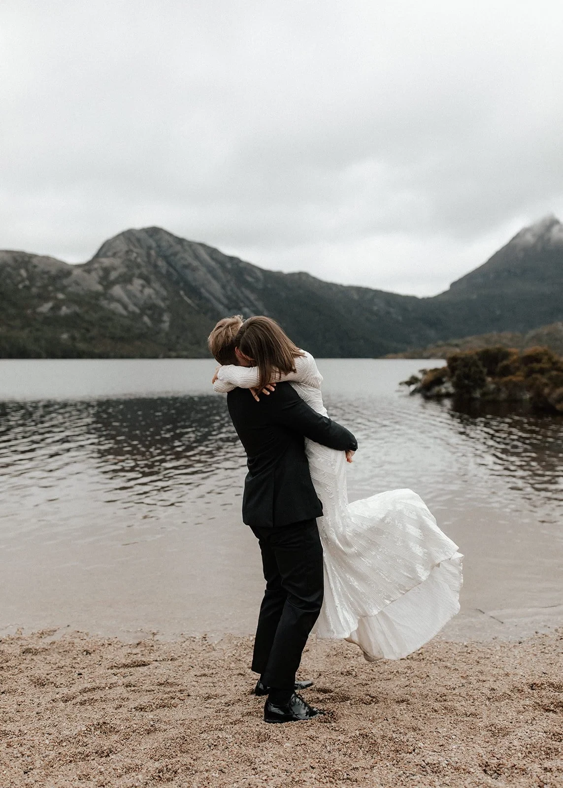 Josh lifting April into an embrace, her dress sweeping above the lakeshore, at Dove Lake, Cradle Mountain, Tasmania, for an Elopement Collective elopement. Photo by Bulb Creative.