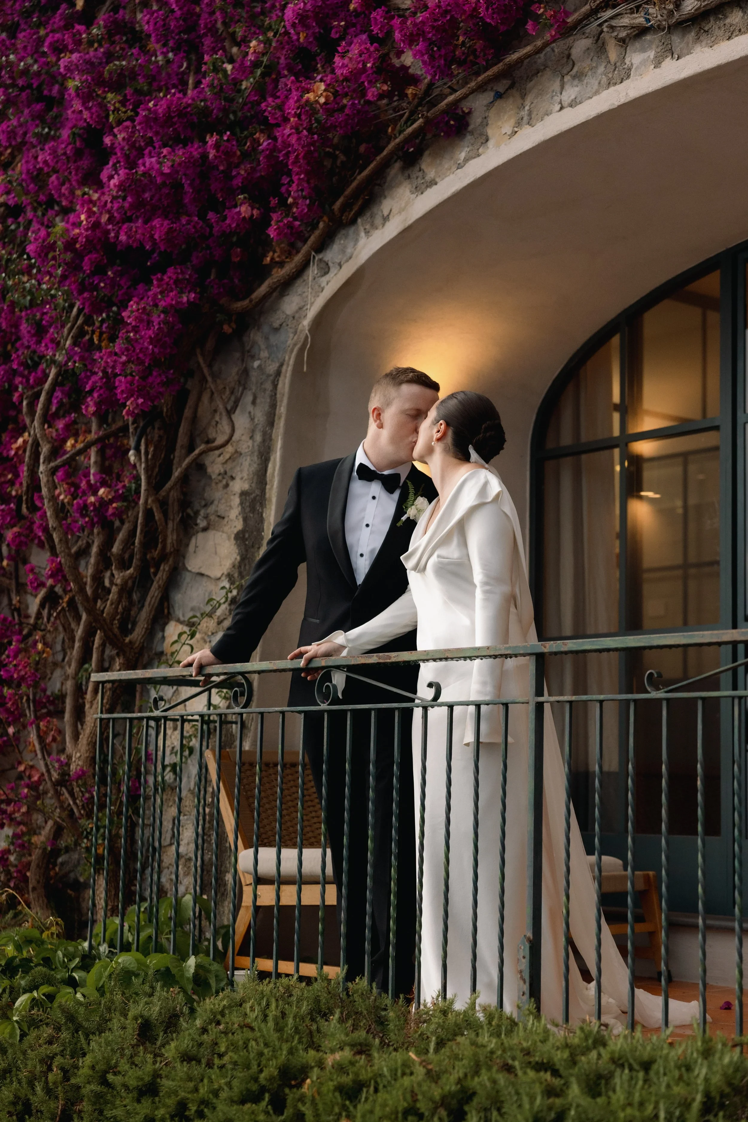 Couple in wedding attire kissing on a balcony framed by flowering plants