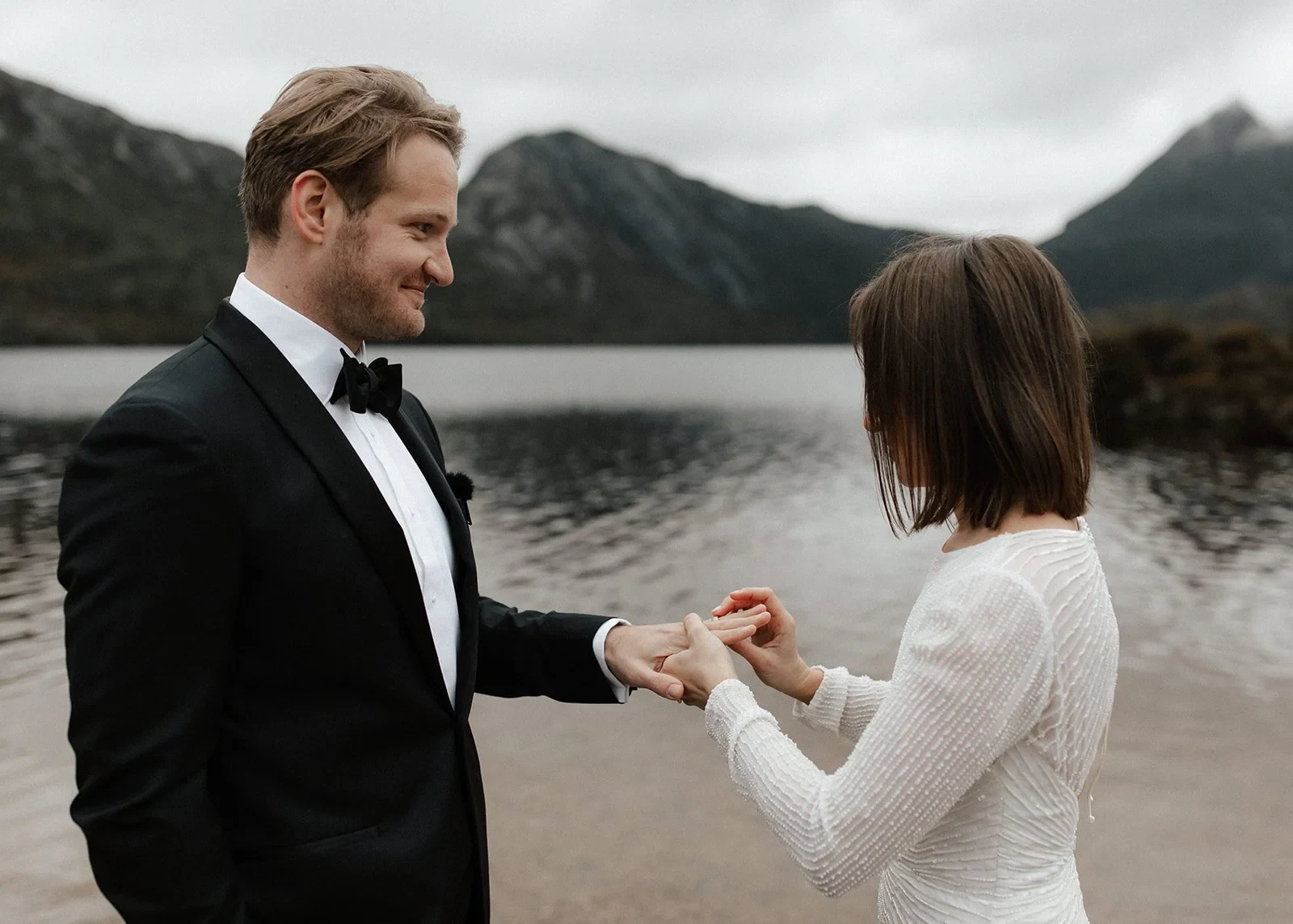 April placing a wedding ring on Josh’s hand beside the lake, at Dove Lake, Cradle Mountain, Tasmania, for an Elopement Collective elopement. Photo by Bulb Creative.