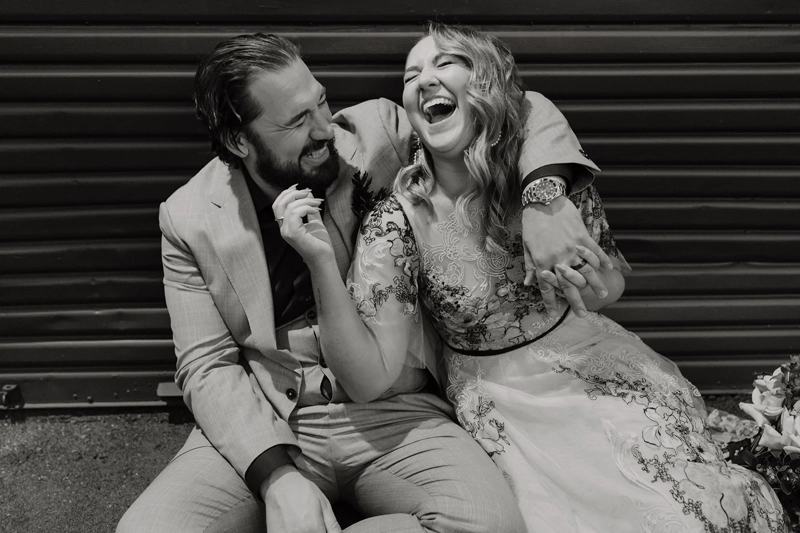 Black-and-white portrait of Victoria laughing with Daniel’s arm around her shoulders while seated outdoors, in Fortitude Valley, Brisbane, for an Elopement Collective elopement. Photo by Tessa Shannon.