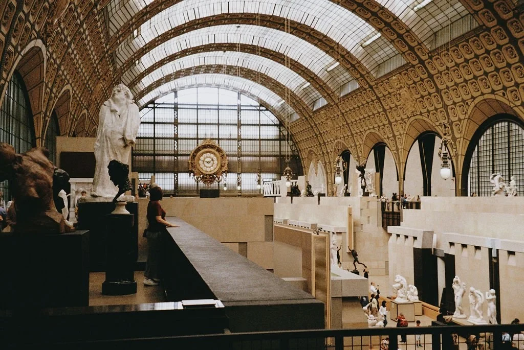 Interior of a museum with large statues and a grand clock, featuring a vaulted glass ceiling and arched windows.