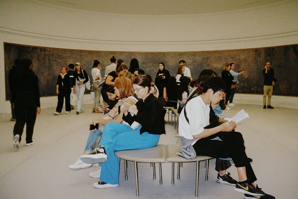 People in a museum room with large paintings, some seated on circular benches, others standing and viewing the art.