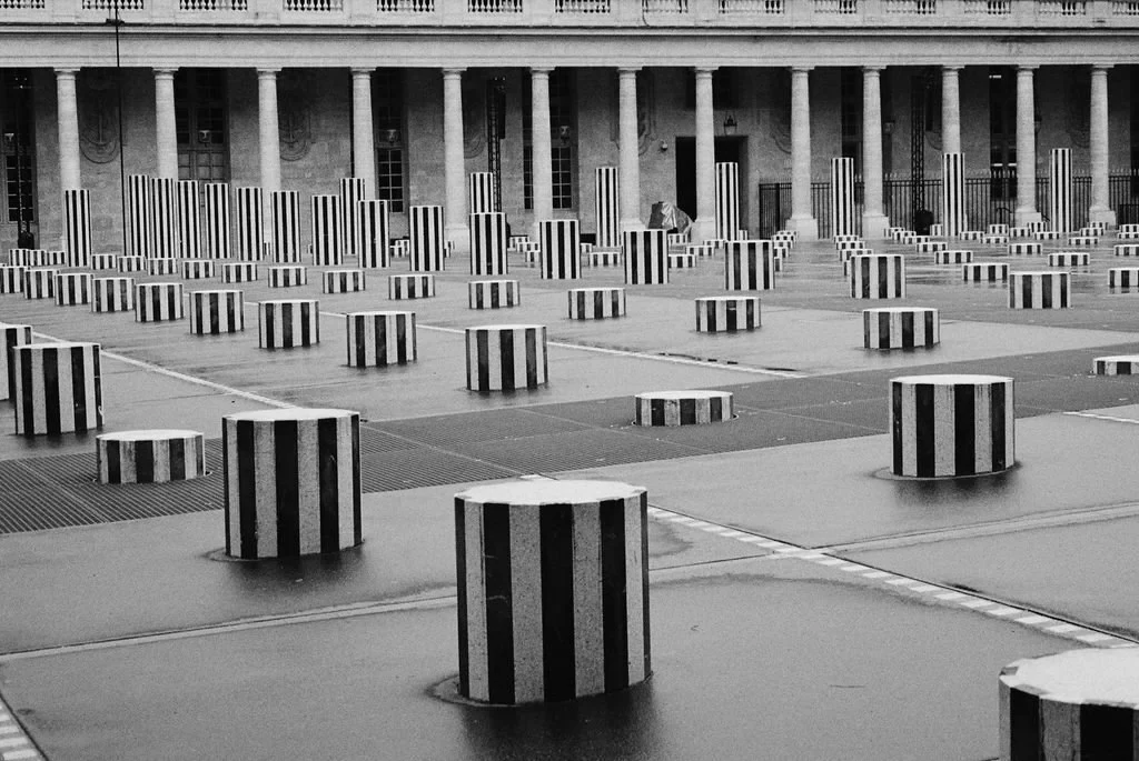 Black and white photo of striped pillars in a courtyard with classical columns in the background.
