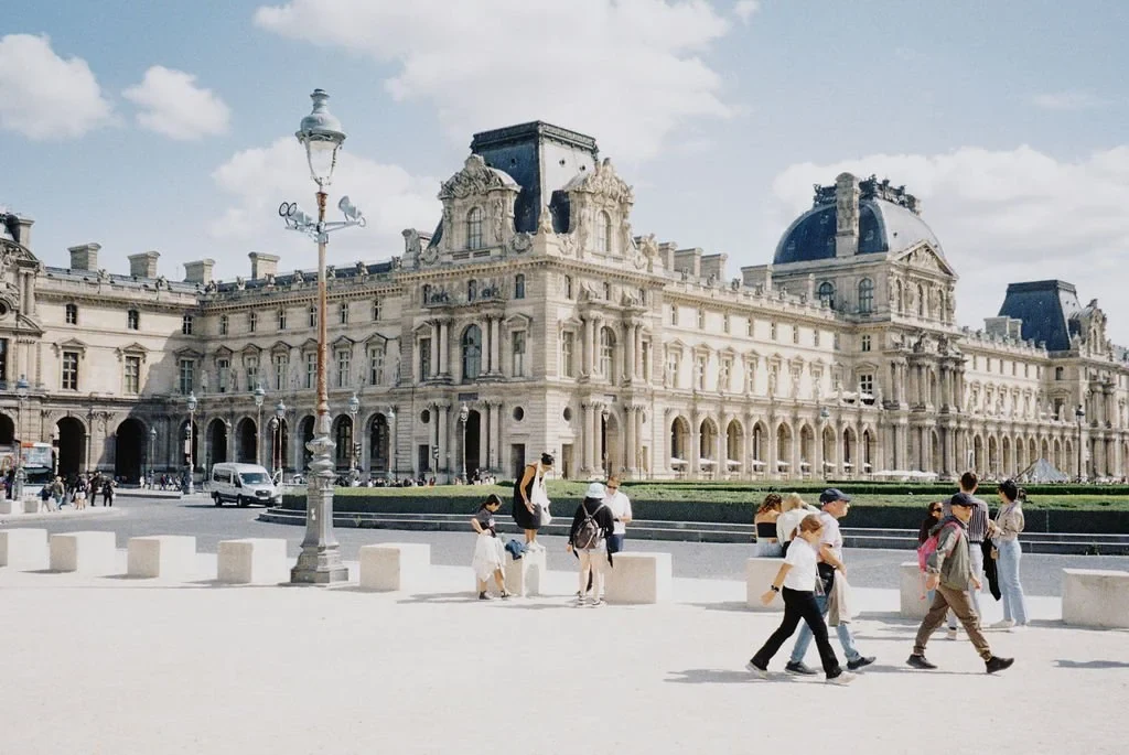 People walking in front of the Louvre Museum in Paris, showcasing its historic architecture under a blue sky.