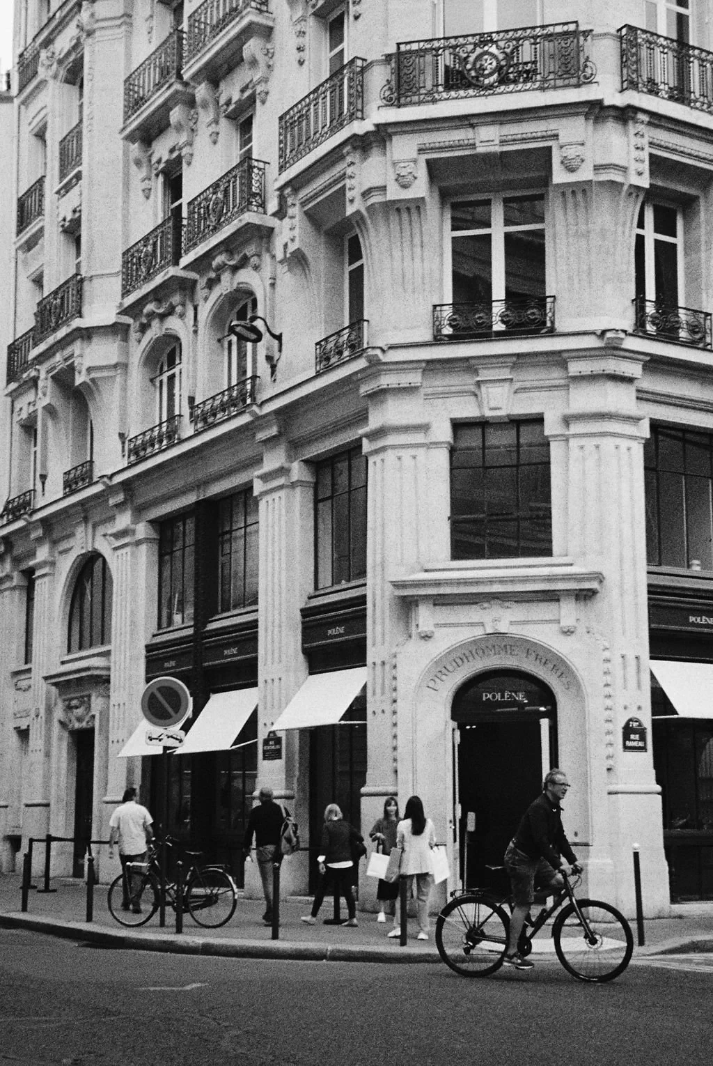 Black and white photo of a street corner with an ornate building, pedestrians, and a cyclist.