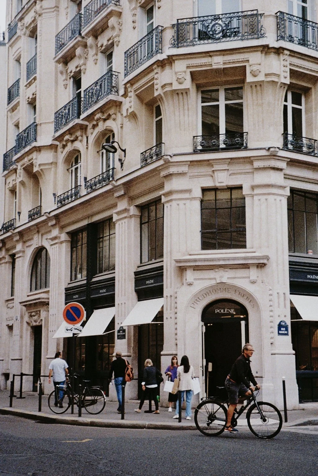 Street view of a historic building featuring ornate architecture, with people walking and a cyclist passing by. The building displays the sign 'PRUDHOMME FRÈRES' above the entrance.