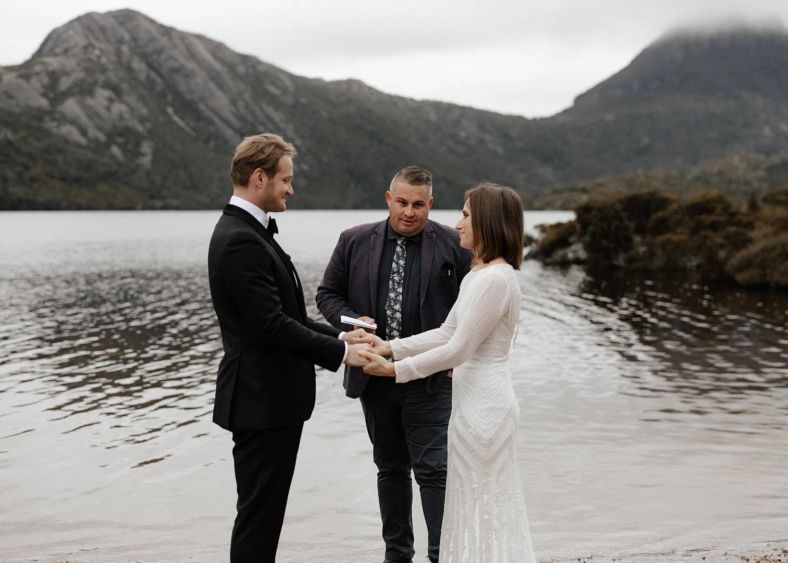April and Josh holding hands during their ceremony with celebrant Josh Withers, at Dove Lake, Cradle Mountain, Tasmania, for an Elopement Collective elopement. Photo by Bulb Creative.