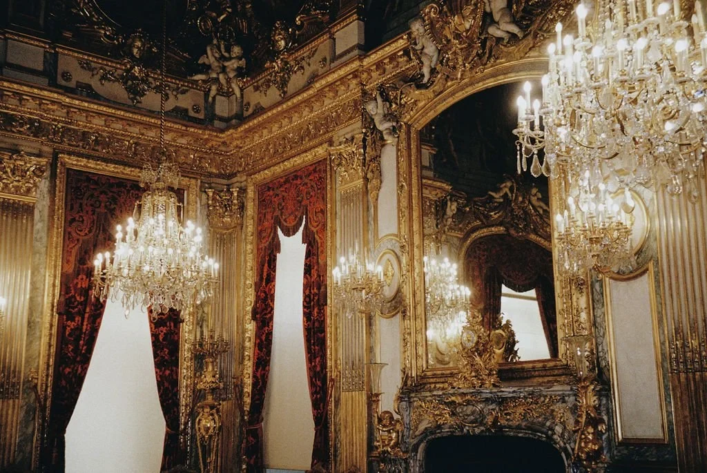 Lavish room with ornate gold decorations, large mirrors, crystal chandeliers, and red velvet drapes.