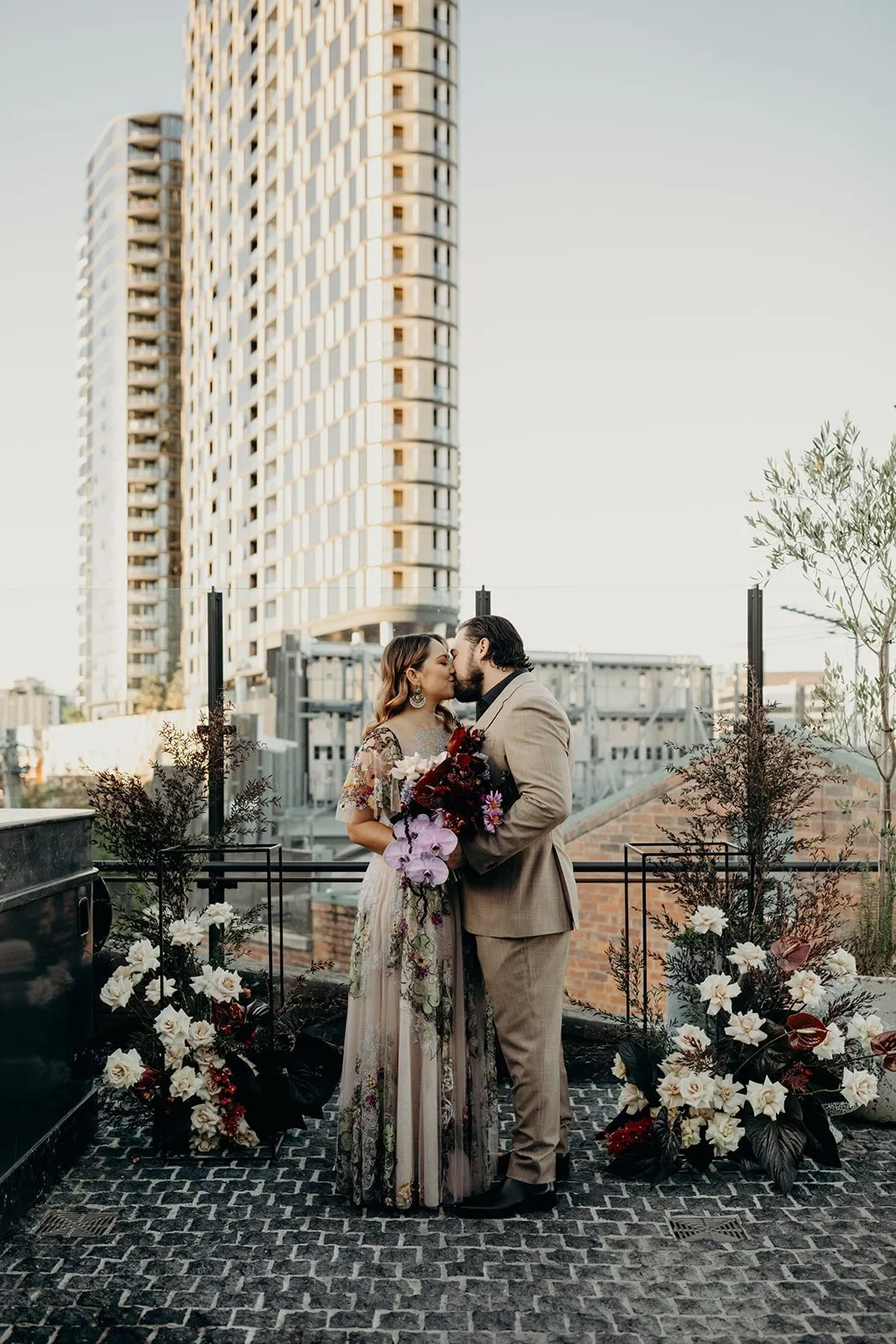 Victoria and Daniel embracing among rooftop ceremony flowers by Mrs Gibbons Flowers, at Agnes Restaurant in Fortitude Valley, Brisbane, for an Elopement Collective elopement. Photo by Tessa Shannon.