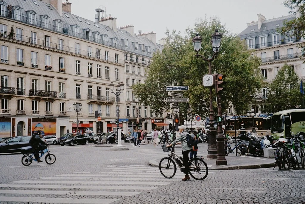 Urban scene with people on bicycles at a city intersection, surrounded by historic European buildings, with cars and trees in the background.
