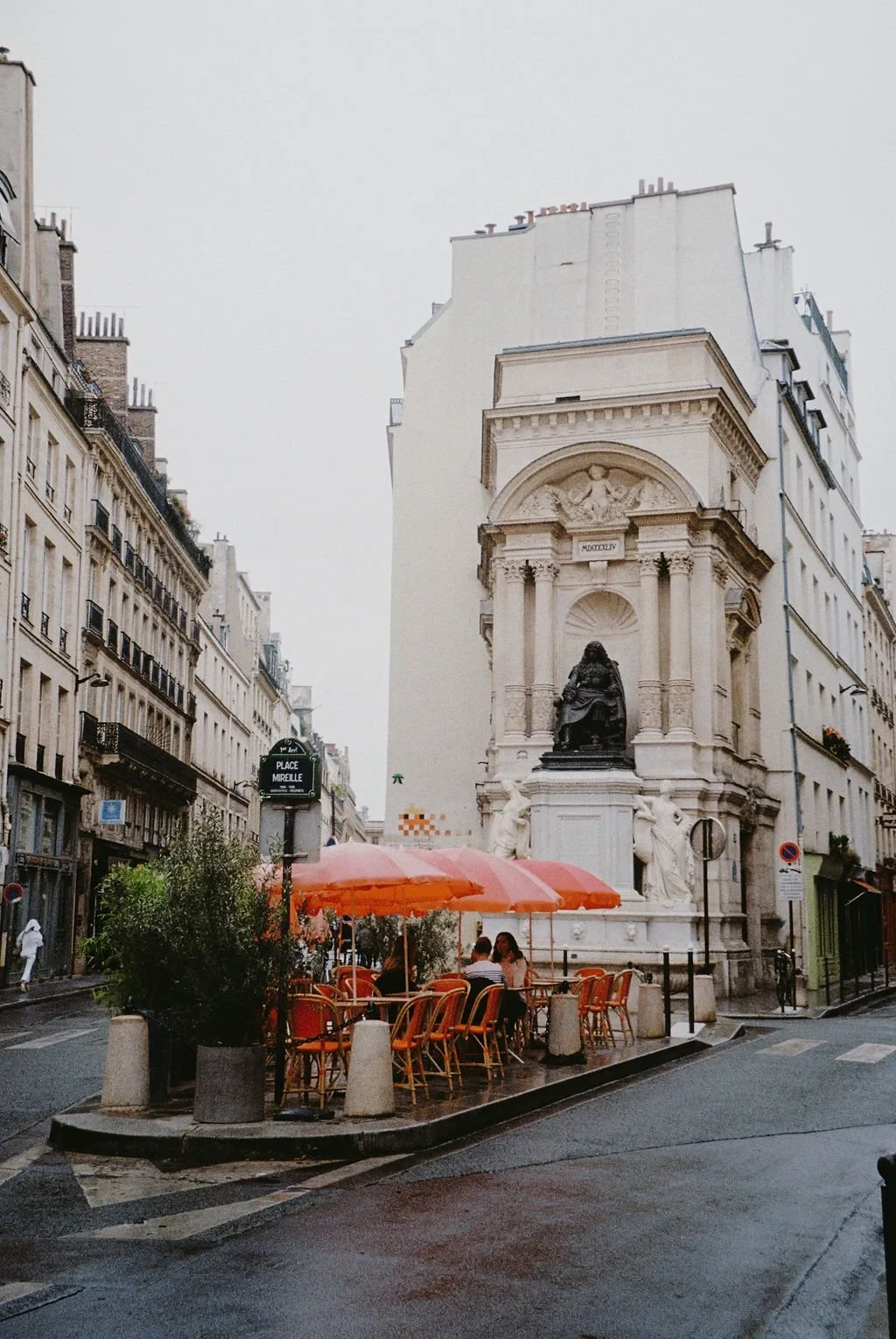 Street scene in Paris featuring a fountain monument, sidewalk café with red umbrellas, and classic Parisian architecture.