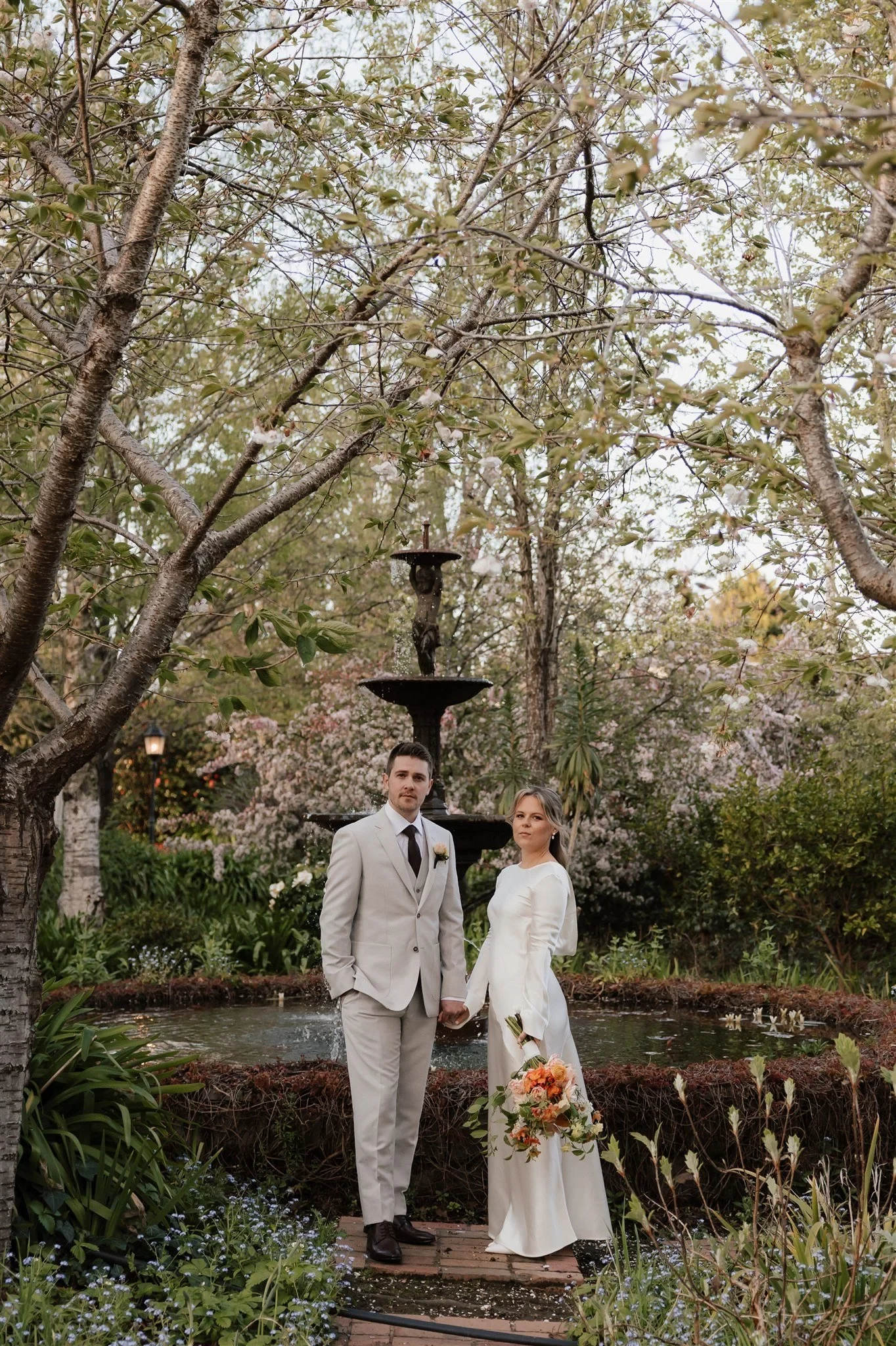 Maddy and Cameron standing hand in hand on the garden path with her bouquet by The Flower Jar and a fountain behind them, at The Mayor’s Residence in Daylesford, Victoria, for an Elopement Collective elopement. Photo by Bulb Creative.