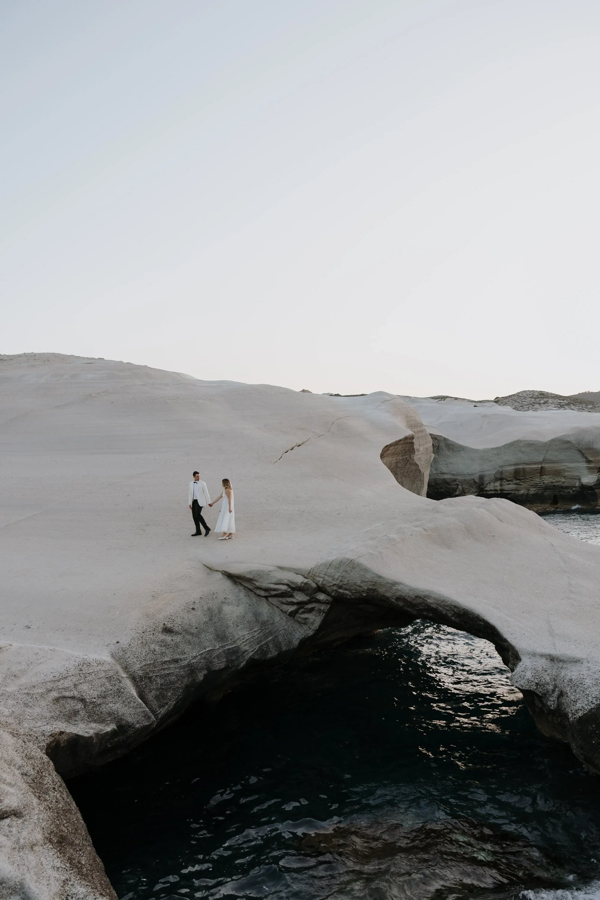 Eloping at Sarakiniko Beach with the Elopement Collective on the Greek Isle of Milos.  A couple is walking across a coastal rock formation with more rocky clifffs in the background.