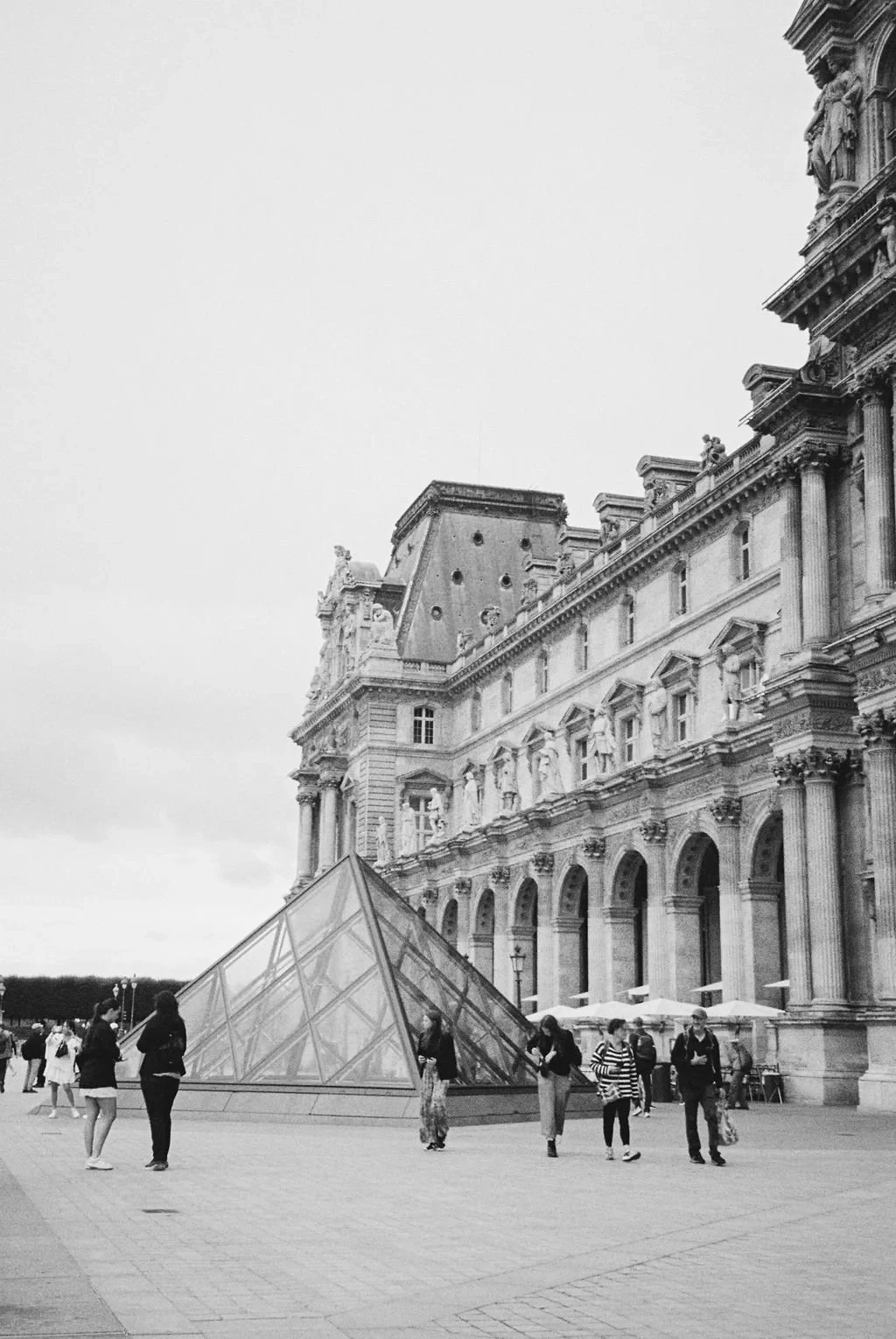 Black and white photo of the Louvre Museum and glass pyramid entrance with tourists walking around.