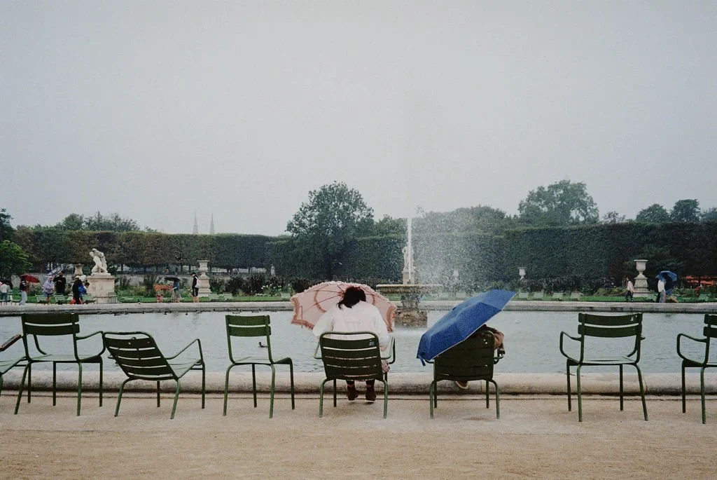 People sitting on green chairs by a fountain in a garden, holding umbrellas on a rainy day.